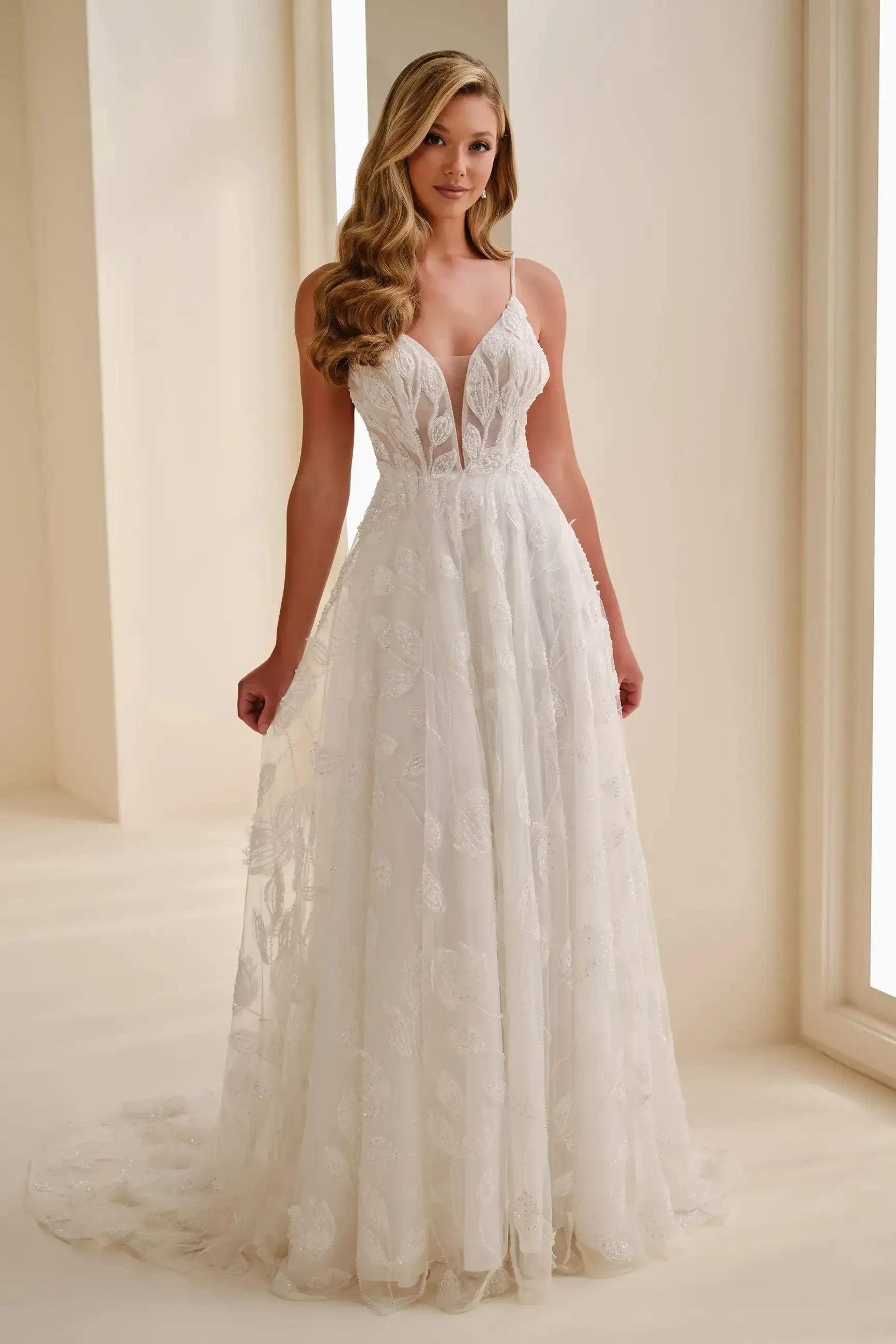 A woman wearing a beautiful white wedding dress with a deep V neckline and delicate floral details, standing in a bright, elegant space.