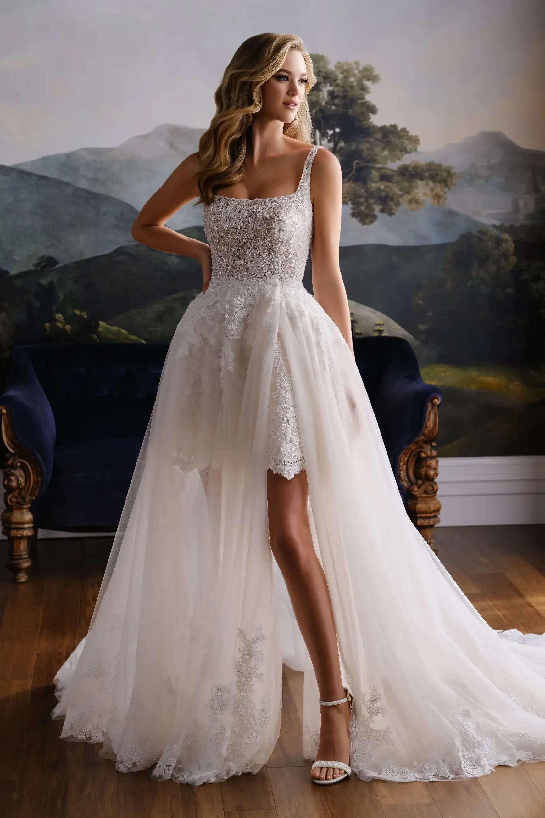 A woman in a bridal gown with a fitted bodice and flowing skirt, standing gracefully against a scenic backdrop. The dress features lace details and has a short front with a longer train at the back. She is wearing strappy heels.